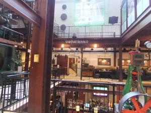Steel I beam in the foreground. The picture looks across the interior of the building into 3 floors of the plant, each surrounded by iron railings. The lower floor shows liquor bottles in the bar. The middle floor shows restaurant tables and booths. The top floor view is of a white wall with 3 dark, round objects on the left and a dark plaque on the right. 