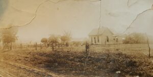 cracked and torn brown tinted photo of old farmstead. A dirt road is in the foreground. A clapboard white house sits behind a wire fence on the right side of the photo. To the left are a few scattered trees in a farmyard.
