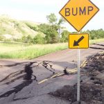 Bump Road sign next to broken and heaved pavement.