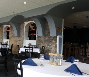 Empty restaurant tables covered with white table clothes, set with silverware and blue cloth napkin tents.